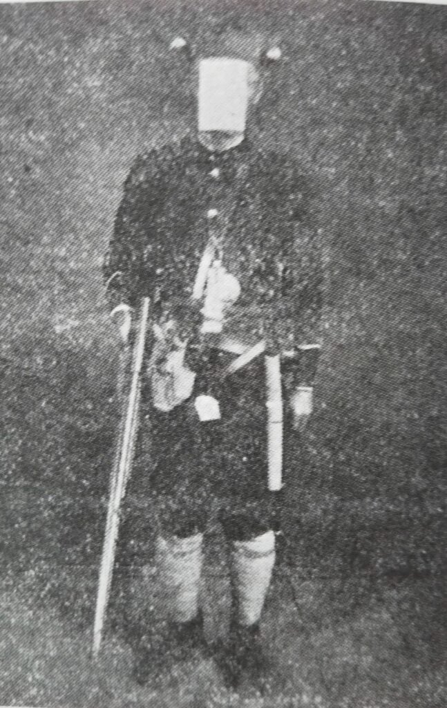 A man dressed to resemble the perpetrator of the Tsuyama Massacre holds a rifle and wears a black school outfit, with numerous weapons strapped to his sides, and two flashlights affixed to his head. 