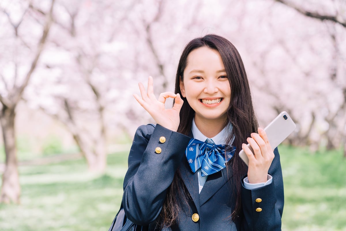Woman with a phone in front of sakura trees holding up an OK sign with her fingers