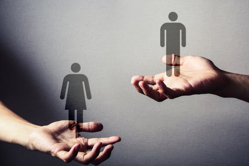 Two hands holding male and female figures, with the female figure held lower to signal inequality