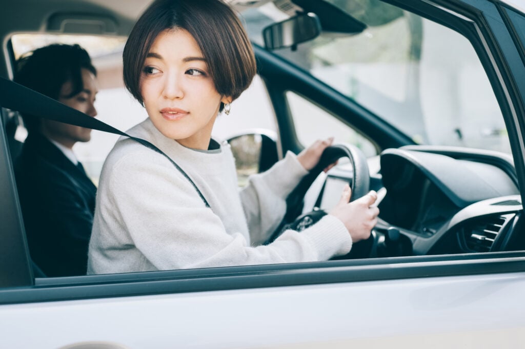 Woman looking out a Japanese driver's side window (right side of the car) while an instructor grades her driving from the left seat