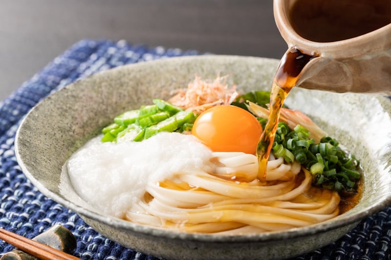 Bowl of udon with an egg in top and udon broth being poured into it