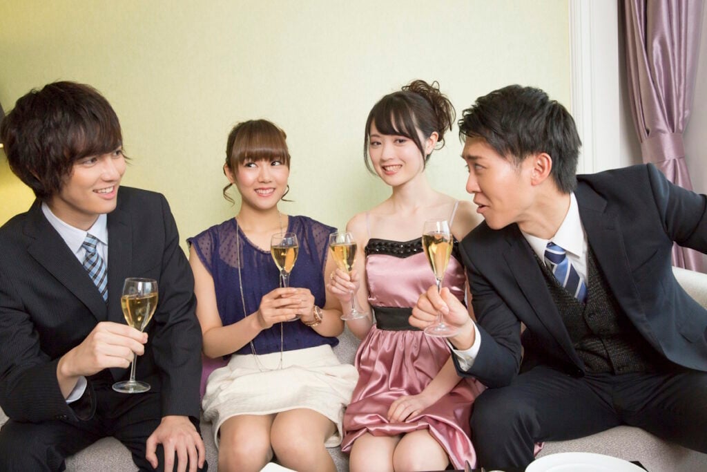 Two men and two women in formal dress holding champagne glasses and talking. The men are talking past the women, who look like they're being paid to smile
