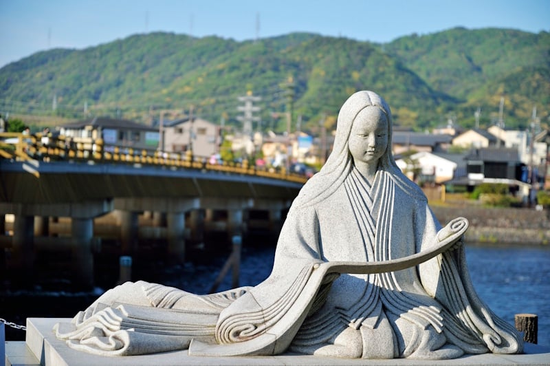 Stone statue of Heian-era author Murasaki Shikibu seated in flowing robes near Uji Bridge, with the river and hills behind