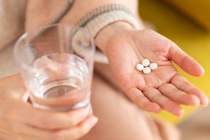 Person in a sweater holding several small white pills in one open palm and a glass of water in the other hand.